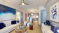 This single-floor residence showcases a thoughtfully designed open-concept layout that seamlessly connects the kitchen and living areas. The kitchen is a standout feature, outfitted with floor-to-ceiling gray cabinetry offering generous storage, stainless steel appliances including a range, built-in microwave, and dishwasher, and a white subway tile backsplash. A central island with a bright white countertop serves as both a prep space and casual dining area, accommodated by modern metal-framed barstools with white cushioned seats. Three cylindrical pendant lights hang above the island, while adjustable track lighting illuminates the perimeter cabinetry. Light wood-tone flooring flows throughout the space, complemented by an area rug beneath the kitchen zone. The open living area features a ceiling fan with integrated lighting and connects directly to the kitchen, creating an airy, cohesive atmosphere. Sliding barn-style doors provide access to adjacent rooms, maintaining the clean, contemporary aesthetic. The overall design emphasizes neutral tones—soft grays, whites, and warm wood accents—resulting in a polished, modern interior that balances functionality with understated elegance.