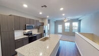 Located on S Wren Dr in Gilbert, Arizona, this single-story home offers a thoughtfully designed open-concept layout that seamlessly connects the kitchen and living areas. The kitchen is a standout feature, showcasing a large granite-topped island with an undermount sink, dark wood cabinetry extending to the ceiling for ample storage, a subway tile backsplash, and a full suite of stainless steel appliances including a French door refrigerator, electric range with microwave above, and a dedicated pantry closet. Wood-look flooring runs throughout the main living and kitchen areas, transitioning to plush carpet in the living room and bedrooms. The living area benefits from natural light streaming through multiple windows and a glass door that opens to the exterior, complemented by a ceiling fan overhead. Bedrooms are carpeted and finished with tray ceilings and ceiling fans, offering a calm and comfortable retreat. The bathroom is well-appointed with a combination tub and shower, granite vanity, wood-look flooring, and direct access to a walk-in closet. A dedicated laundry room houses a full-size washer and dryer with an overhead shelf for added convenience. Neutral gray tones throughout the home create a clean, modern aesthetic.