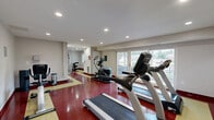 Located at 3302 Lockheed Blvd in Alexandria, Virginia, this fitness center is a well-appointed, single-floor facility divided into two functional workout areas. The main cardio room is bright and open, benefiting from large floor-to-ceiling windows that invite natural light and offer views of the surrounding neighborhood greenery. This area is equipped with multiple treadmills and an elliptical trainer, all positioned to take advantage of the outdoor views. The flooring throughout features a bold, textured pattern in red and yellow, lending the space a dynamic and energetic aesthetic. Recessed lighting in the white ceiling ensures consistent illumination throughout. A full-length mirror in one section visually expands the space and supports workout form checks. The second room is dedicated to strength training, housing a multi-station cable and weight machine along with free weights and a hyperextension bench, making it suitable for a comprehensive resistance training routine. A window in this room provides additional natural light. A clean, tiled shower/bath area is also present, providing a convenient post-workout rinse facility. Informational notices are posted near the entry, and a hand sanitizer dispenser reflects attention to hygiene standards. Together, the two rooms form a compact yet comprehensive fitness facility.