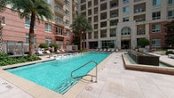 This pool area and adjacent lobby corridor offer a well-appointed amenity space within a high-rise residential community. The outdoor pool is a centerpiece feature, showcasing clear turquoise water framed by a broad, light-toned tile deck. A row of lounge chairs lines the perimeter, providing ample space for relaxation. Towering palm trees add a resort-like atmosphere, while the surrounding multi-story buildings with balconies create a sheltered, private courtyard setting. A spa or water feature sits adjacent to the main pool, enhancing the overall recreational appeal.

Access to the interior is provided through a secure gate and a set of glass double doors, leading into a refined lobby corridor. This transitional space features neutral-toned walls, recessed lighting, and wall-mounted sconces that cast a warm, welcoming glow. The flooring transitions between light tile and patterned carpet runners, adding visual interest. Columns punctuate the corridor, lending an architectural formality to the passage. Curated abstract artwork adorns the walls, contributing an upscale aesthetic. Seating with decorative pillows is positioned along the corridor, offering a comfortable waiting area. Views through the corridor's end windows frame the pool courtyard, maintaining a visual connection between interior and exterior spaces.