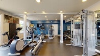 Located at 5680 Bay St in Emeryville, California, this well-equipped fitness center offers a comprehensive workout environment within a compact, single-floor layout. The space features durable rubber flooring throughout, recessed ceiling lighting, and a bold deep-blue accent wall that anchors the cardio zone, where a wall-mounted flat-screen TV provides entertainment during workouts. The cardio section is stocked with treadmills, elliptical trainers, a rowing machine, and a stationary bike, catering to a wide range of aerobic exercise preferences. A dual-cable functional trainer serves as the centerpiece for strength training, complemented by a full rack of dumbbells neatly organized along the mirrored wall. Floor-to-ceiling mirrors amplify the sense of space and allow users to monitor their form throughout their sessions. A dedicated functional training zone features TRX suspension straps and wall-mounted medicine balls, supporting bodyweight and resistance-based workouts. Large sliding glass doors flood the space with natural light and open directly to an outdoor patio, offering a seamless connection to the exterior. Structural columns are integrated into the layout without impeding movement, and the overall design balances practicality with a clean, modern aesthetic suited to everyday fitness use.