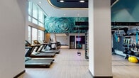 This contemporary fitness center is thoughtfully designed to deliver a premium workout experience. The space is anchored by soaring double-height ceilings and floor-to-ceiling windows that flood the interior with abundant natural light, offering views of the surrounding outdoor greenery. Light wood-style flooring runs throughout, complementing the clean, modern aesthetic.

A row of Matrix treadmills and elliptical machines lines the windowed perimeter, providing an energizing environment for cardio workouts. Adjacent to the cardio zone, a dedicated functional training area features a RAX multi-functional rig loaded with TRX suspension trainers, gymnastic rings, and resistance attachments. A Matrix adjustable bench and a comprehensive free weights section — including a full rack of kettlebells and medicine balls — support strength and conditioning routines.

A standout feature of the space is a large-scale swirling teal mural that wraps dramatically across both the wall and ceiling, creating a bold artistic focal point. A neon "BEAST MODE" sign illuminates a built-in amenity station equipped with a water filtration unit, storage shelving, and personal lockers — adding both function and personality to the space. Recessed and cove lighting throughout enhances the dynamic, motivating atmosphere of this well-appointed fitness facility.