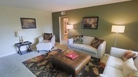 This single-story space showcases a well-appointed living room that serves as the focal point of the home. The room is anchored by a bold olive-green accent wall, which creates a warm and inviting atmosphere that contrasts with the lighter neutral walls on the remaining sides. The textured ceiling adds architectural character overhead, while carpeted flooring runs throughout, adding to the overall sense of comfort.

The living area is furnished with a light gray sofa accented by earth-toned throw pillows, complemented by a matching armchair and ottoman arrangement. A large tufted leather ottoman serves as a central coffee table, grounding the seating arrangement atop a richly patterned area rug featuring deep reds, greens, and golds. Framed artwork on the walls adds a finishing touch to the curated aesthetic.

A doorway visible from the living room leads to an adjacent space, suggesting an open and connected layout. A floor lamp provides warm supplemental lighting, enhancing the cozy ambiance. The overall design palette of greens, browns, and neutrals gives the space a cohesive, nature-inspired feel throughout.
