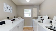 The Clothes Care Center is a single-floor laundry facility designed with functionality and convenience in mind. The room is finished with neutral gray walls and durable tile flooring, creating a clean and practical environment. Multiple top-loading washing machines and dryers are arranged along opposing walls, maximizing the use of available space and allowing several users to do laundry simultaneously. A centrally positioned window on the back wall allows natural light to fill the room, contributing to a bright and open atmosphere. Overhead, a flush-mounted ceiling light provides consistent illumination throughout. An electrical panel is mounted on the wall near the window, keeping utilities accessible and organized. The overall layout is straightforward and efficient, with each machine positioned for easy access. The facility is well-maintained, with white appliances standing out against the light-toned walls and flooring. This clothes care center serves as a practical shared resource, equipped to handle laundry needs with multiple machines available for simultaneous use.