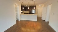 This single-story space showcases an open-concept layout that seamlessly connects the living area to the kitchen. The kitchen is a standout feature, equipped with dark espresso cabinetry that provides a striking contrast against the light granite countertops. Stainless steel appliances, including a refrigerator and built-in microwave, add a modern, polished touch. A peninsula with a sink serves as both a functional workspace and a natural dividing element between the kitchen and living area, ideal for casual dining or entertaining.

Three pendant lights hang above the peninsula, complementing the track lighting installed along the kitchen ceiling, ensuring the space is well-lit throughout. Rich, warm-toned hardwood flooring extends across the entire open area, adding depth and cohesion to the design.

A built-in shelving unit is recessed into the wall adjacent to the kitchen, offering practical display and storage space. The neutral wall tones enhance the overall brightness of the interior. A hallway visible near a white paneled door suggests additional rooms beyond the main living area, contributing to the functional flow of the floor plan.