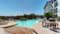 This single-story amenity space offers a well-appointed combination of outdoor recreation and refined interior gathering areas. The centerpiece of the outdoor area is a generously sized, freeform swimming pool with clear turquoise water, complemented by an adjacent spa with water features. The pool deck is finished in warm, irregular flagstone pavers that add texture and a natural aesthetic. Surrounding the pool are multiple lounge chairs and patio seating arrangements, including a thatched umbrella shade structure, providing ample space for relaxation. The area is enclosed by a wrought iron fence and accented with lush greenery, offering a sense of privacy while maintaining open views of the surrounding urban landscape under expansive open skies.

Inside, the interior spaces are finished with rich, dark wood trim, panel doors, and wood-framed windows fitted with horizontal blinds. Hallways are clean and well-lit, leading to multiple rooms. A formal conference or meeting room features a substantial solid wood table paired with upholstered striped chairs beneath a wrought iron chandelier with cream glass shades. Warm terracotta-toned walls, decorative crown molding with wood beam accents, and curated artwork contribute to a cohesive, inviting atmosphere throughout the interior.