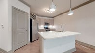 Located on Deer Dr in the desirable Lakewood Ranch community of Sarasota County, Florida, this single-floor unit offers a thoughtfully designed open-concept layout with modern finishes throughout. The kitchen is a standout feature, showcasing a large white quartz island with an integrated sink, stainless steel appliances including a refrigerator, range, and microwave, and two-tone cabinetry with warm wood-grain uppers accented by a decorative lattice detail. Stylish pendant lights and a track lighting fixture illuminate the space, which flows seamlessly into the expansive living area. The living room features light wood-style plank flooring, crown molding, a ceiling fan, and natural light from windows fitted with horizontal blinds, along with direct access to an exterior door. Bedrooms are finished with plush carpeting, crown molding, and generous proportions, providing a comfortable and quiet retreat. The bathroom is clean and contemporary, featuring wood-toned cabinetry, a framed mirror, a well-lit vanity, and light plank flooring. Throughout the unit, a neutral color palette of whites and soft greiges creates a bright, cohesive aesthetic that is both modern and welcoming.