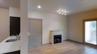 This single-floor space presents a clean, contemporary design with a neutral color palette that creates a bright and welcoming atmosphere. The main living area is immediately notable for its light-toned hardwood flooring, which runs throughout and lends warmth and continuity to the space. A built-in fireplace with a structured surround serves as a central focal point, offering both aesthetic appeal and functional comfort. The fireplace mantel features a simple, modern design that complements the minimalist aesthetic of the room. Recessed ceiling lighting and a sleek multi-bulb track light fixture provide ample, well-distributed illumination throughout the area. The entryway is framed by a solid, paneled door finished in a soft gray tone, consistent with the overall color scheme. Smooth, light-beige walls and clean baseboards contribute to the polished, move-in-ready feel of the unit. The open layout allows for flexible furniture arrangement, making the living area adaptable to various lifestyle needs. Overall, the space combines functional design with understated modern finishes to deliver a comfortable and practical living environment.