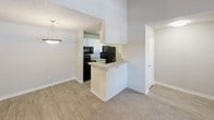 This single-story unit offers a practical and well-organized layout with a neutral color palette throughout. The living room serves as the focal point, featuring plush wall-to-wall carpeting, a ceiling fan, a wood-burning fireplace framed in gray tile, and dual windows alongside a secondary door that allow ample natural light to fill the space. Textured ceilings are present throughout the unit, adding a subtle architectural detail.

The kitchen is efficiently designed with white cabinetry, a subway tile backsplash, a breakfast bar peninsula, and black appliances including a refrigerator, range, and microwave. Wood-look vinyl flooring runs through the kitchen and connects to the adjacent dining area, which is accented by a hanging pendant light. The transition between the kitchen and living areas is defined by a partial wall, maintaining an open yet structured feel.

The bedroom is generously proportioned, featuring soft carpeting, a large window with blinds, and ample wall space. The bathroom includes wood-look vinyl flooring, a vanity with a mirrored medicine cabinet, and a full tub and shower combination with tiled surround and brushed nickel fixtures. A closet is conveniently accessible from the bathroom hallway, providing practical storage within the unit.