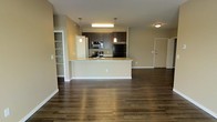 This single-floor space showcases an open-concept layout that seamlessly connects the kitchen and living areas, creating a spacious and functional environment. The kitchen is a standout feature, outfitted with dark espresso cabinetry that extends to the upper walls, stainless steel appliances including a refrigerator, microwave, and range, and a peninsula-style island topped with light stone countertops. Pendant lights hang above the island, providing focused task lighting while adding a modern aesthetic touch. Rich, dark hardwood flooring runs throughout the space, offering a warm contrast to the neutral beige walls and white trim. A built-in shelving unit is visible along one wall, offering practical storage and display options. The open layout allows for flexible furniture arrangements in the living area, while the clean sightlines between spaces contribute to an airy, well-connected feel. Additional doors suggest access to further rooms or storage beyond the main living space. Overall, the design balances contemporary finishes with practical functionality, making it a well-appointed and visually cohesive space.