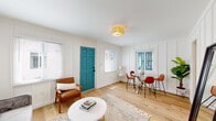 This thoughtfully designed single-floor home blends classic charm with modern updates throughout. The living area welcomes visitors with crisp white board-and-batten wall paneling, warm light hardwood floors, and abundant natural light streaming through multiple windows fitted with white blinds. A bold teal front door serves as a striking focal point, adding a playful pop of color against the all-white interior. A woven pendant light fixture overhead completes the inviting atmosphere.

The adjacent dining nook features a glass-top table with cognac leather chairs, framed by a paneled accent wall and a bright window with greenery peeking through. The galley-style kitchen is a standout, showcasing white Shaker cabinetry, white quartz countertops, a farmhouse sink, a subtle tile backsplash, and a set of retro-style teal appliances — including a matching refrigerator, range, and hood — that infuse the space with personality.

A utility room with ample white cabinetry and countertop workspace provides excellent storage and function. The bathroom offers a serene retreat with a white freestanding vanity, polished nickel fixtures, a medicine cabinet mirror, and intricate patterned tile flooring. An additional versatile room with clean white walls and hardwood floors rounds out the layout, ideal as a bedroom or office.