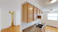 This single-floor residence offers a thoughtfully designed layout with a clean, functional aesthetic. The kitchen serves as a central feature, showcasing warm honey-toned wood cabinetry that provides generous storage space. The upper cabinets are positioned prominently, offering ample room for organization, while the lower cabinetry complements the overall design seamlessly. A double-basin stainless steel sink is set into granite-style countertops that add a touch of understated elegance to the space. The kitchen opens to adjacent areas, creating a connected and accessible flow throughout the home. Hardwood flooring extends from the kitchen into the surrounding rooms, adding warmth and continuity to the interior. The walls are finished in crisp white, enhancing the sense of brightness and space throughout. Natural light filters into the rooms, contributing to a welcoming ambiance. The open passageway visible from the kitchen connects to additional rooms, suggesting a practical and well-organized floor plan. Overall, this space combines classic wood finishes with clean architectural lines to create a comfortable and functional single-floor living environment.