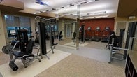 This fitness center is designed to support a comprehensive workout experience within a single, open-plan floor. The space is thoughtfully organized into distinct functional zones, separating strength training equipment from cardiovascular machines. The strength training area features a selection of cable and resistance machines, providing users with a variety of muscle-targeting options. Adjacent to this zone, a row of treadmills lines the far wall, offering ample cardio options for users of all fitness levels.

Large, floor-to-ceiling mirrors are strategically positioned throughout the space, visually expanding the room and allowing users to monitor their form during exercise. The flooring transitions between durable, cushioned rubber matting in the equipment zones and a neutral carpet area, offering both safety underfoot and comfort. Overhead, bright fluorescent strip lighting ensures the entire space is well-illuminated, creating a clean and energizing atmosphere.

The walls feature a warm, earthy color palette with deep red and neutral tones, contributing to a motivating ambiance. The layout allows for generous spacing between machines, ensuring ease of movement and accessibility throughout the fitness center.