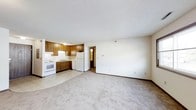 This single-floor space features an open-concept layout that seamlessly connects the living area and kitchen, creating a functional and cohesive environment. The kitchen is equipped with warm wood-toned cabinetry offering generous storage, white appliances including a refrigerator, range, and dishwasher, and a countertop with an integrated sink. The cabinetry extends to the ceiling, maximizing vertical storage space. A textured white ceiling runs throughout the space, complemented by neutral white walls that enhance the sense of openness and light. The living area is finished with wall-to-wall carpeting in a neutral tone, providing a comfortable and practical underfoot surface. The transition between the kitchen's hard flooring and the carpeted living area is clearly defined, adding visual structure to the open floor plan. A hallway visible in the background suggests access to additional rooms, offering further utility beyond the main living space. The overall ambiance is clean, straightforward, and practical, with a design that prioritizes functionality and ease of use. The neutral color palette throughout creates a versatile backdrop well-suited to a variety of furnishing styles.