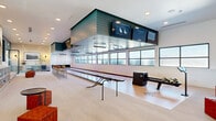 This generously sized single-floor bowling lounge and entertainment venue offers an upscale recreational experience defined by thoughtful design and premium amenities. The bowling area features polished full-length lanes illuminated by UV accent lighting and overhead lane monitors mounted beneath a striking dark-paneled soffit structure. A row of high-top bar seating runs alongside the lanes, providing an ideal vantage point for spectators, while ball return equipment and bowling ball storage cubbies are neatly integrated into the layout.

The adjoining lounge area is finished in a contemporary palette of warm neutrals and deep teal accent walls, creating an inviting and modern atmosphere. Built-in bench seating with plush cushions lines the walls, complemented by clusters of terracotta woven ottomans and sleek round side tables. A centrally featured arched display niche with curated artwork serves as a focal point, surrounded by framed landscape prints and warm pendant lighting along the perimeter walls.

Large gridded windows allow diffused natural light to fill the space, enhancing the open and airy ambiance throughout. Recessed ceiling lighting provides consistent illumination across the expansive floor plan. The space is well-suited for both casual recreational use and private social gatherings, blending entertainment functionality with refined interior design.