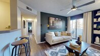 This thoughtfully designed single-floor residence makes a strong visual statement through its consistent use of deep teal accent walls, warm wood-style flooring, and contemporary finishes. The living room serves as the heart of the home, anchored by a striking teal feature wall adorned with framed artwork and complemented by a ceiling fan, built-in shelving, and sliding glass doors that open to an outdoor balcony, inviting natural light throughout. A dedicated dining area near the entry provides a welcoming gathering space, while the galley-style kitchen is equipped with stainless steel appliances, white cabinetry, and ample counter space. A hallway connects the living spaces to the bedrooms, each carrying the signature teal palette. The primary bedroom is spacious and serene, featuring a large tufted headboard wall, a generous window with outdoor views, and access to a private balcony. A secondary bedroom continues the design theme with moody blue-gray walls, natural light, and a cozy layout. Both bathrooms are clean and functional, each featuring a tub-shower combination, wood-style flooring, and neutral finishes with brushed gold hardware accents. The cohesive design language and bold color choices give this residence a distinctive, modern character throughout.