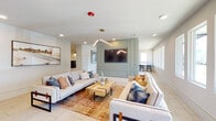 Located at Richmond Row in Suwanee, Georgia, this single-level community amenity space offers a well-appointed and thoughtfully designed environment for residents to gather, relax, and stay active. The entrance greets visitors with striking red double doors, large windows that invite abundant natural light, and clean large-format tile flooring that carries throughout the space. The main lounge area serves as the social heart of the building, featuring two generously sized neutral sofas arranged around a wooden crate-style coffee table atop a warm patterned area rug. A sage green paneled accent wall frames a wall-mounted flat-screen TV, while a modern angular chandelier adds an architectural focal point overhead. Adjacent to the lounge, the dining and gathering room is bathed in natural light from a full row of windows overlooking the neighborhood. A solid wood communal dining table surrounded by mixed seating and two statement gold starburst pendant lights create an inviting, café-like atmosphere. A dedicated coffee bar area showcases teal cabinetry, marble-look countertops, and a bold herringbone tile backsplash — a stylish and functional amenity. The floorplan also incorporates a fitness area, as visible in the dollhouse view. Throughout, the design blends contemporary finishes with warm accents to create a cohesive and welcoming community hub.