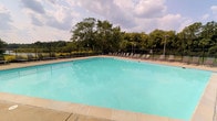 This outdoor recreational space centers around a generously sized swimming pool with clear, turquoise water that reflects the open sky above. The pool is a prominent feature, offering ample room for swimming and leisure. Surrounding the pool is a wide, tiled deck finished in neutral tones, providing plenty of space for relaxation and movement. A row of lounge chairs lines the perimeter of the deck, offering dedicated spots for sunbathing and rest. The entire area is enclosed by a sturdy wrought iron fence, which enhances both safety and visual appeal while maintaining an open, airy feel. Beyond the fence, a backdrop of mature deciduous trees creates a lush, green boundary that adds a natural, park-like ambiance to the setting. Tall light poles are positioned around the area, suggesting the space is equipped for evening use. The overall atmosphere is inviting and well-maintained, combining functional amenities with a pleasant outdoor environment. This pool area is well-suited for both casual recreation and community gatherings, offering a clean and organized outdoor retreat with a serene natural backdrop.