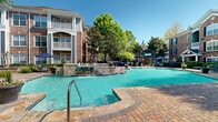 Located at 3001 Northeast Expressway in Atlanta, GA, this outdoor amenity space delivers a resort-style experience within a well-maintained residential community. At its centerpiece sits a generously sized swimming pool with crystal-clear turquoise water, complemented by a separate spa featuring a stone waterfall feature and elegant stonework surround. The pool deck is finished with attractive brick pavers and natural stone accents, offering ample space for relaxation and recreation.

Surrounding the pool, a well-appointed sun deck is lined with blue lounge chairs and shaded by mature deciduous trees, providing a comfortable outdoor retreat. Thoughtfully placed landscape lighting and classic lamp posts add charm to the environment, while manicured shrubs, flowering plants, and large decorative planters contribute to the lush, polished aesthetic throughout.

The adjacent clubhouse is a standout architectural feature, designed in a traditional brick style with arched entryways, columned porticos, a pitched roof, and chimney details that evoke a refined, classic character. An outdoor pergola structure nearby provides additional shaded gathering space, accompanied by an outdoor fireplace and dining areas suited for socializing. The multi-story residential buildings, constructed of warm brick with white-trimmed balconies, frame the amenity area and contribute to the community's cohesive, upscale atmosphere.