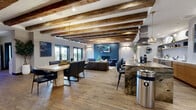 This thoughtfully designed single-floor space seamlessly blends rustic character with modern sophistication across a generously sized open floor plan. The most striking architectural feature is the series of rich, exposed wood beams that span the ceiling throughout, adding warmth and visual depth to the bright, airy interior. Wide-plank hardwood flooring flows continuously underfoot, unifying each area with a cohesive, natural aesthetic.

The open layout encompasses a comfortable living area furnished with a channeled leather sofa and eclectic accent seating, alongside a defined dining space anchored by a substantial table with seating for several guests. Large glass doors along one wall invite natural light deep into the space and provide a direct connection to the outdoors.

The kitchen is a true centerpiece, featuring a dramatic stone island with bold, book-matched veining and an integrated cooktop beneath a stainless steel range hood. Warm wood cabinetry lines the walls alongside open shelving styled with curated décor, and pendant lighting adds an elegant finishing touch. A dedicated display and storage wall near the entry offers additional functional space.

The overall ambiance is warm, inviting, and design-forward — a well-appointed space that balances livability with refined style.