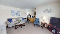 Skytop Gardens offers a thoughtfully arranged single-floor residence designed for comfortable everyday living. The property includes an on-site leasing office, which features a welcoming reception area furnished with a tufted gray sofa, upholstered accent chairs, a glass-top coffee table, and a staffed front desk — creating an inviting first impression for visitors and residents alike.

The living area is carpeted and bathed in neutral tones, with wall-mounted décor and ample space for seating arrangements. A window unit provides climate control, and natural light filters in through a large window dressed with grommet-style curtains.

The bedroom is generously sized, benefiting from multiple windows that allow natural light to fill the room. Dark wood furnishings, including a substantial dresser with mirror, complement the carpeted flooring and create a warm, restful atmosphere.

The kitchen features warm oak cabinetry, white appliances including a range, and a stainless steel sink. A dining nook adjacent to the kitchen is brightened by a window with a floral valance and decorative wall art.

The bathroom includes a tiled tub-shower combination with chrome fixtures, a window for ventilation, and coordinating décor. Throughout the residence, a consistent neutral palette and warm accents contribute to a cozy, lived-in ambiance.