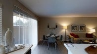 This single-floor space showcases an open-concept layout that seamlessly connects the living and dining areas, creating a cohesive and functional environment. The dining area is anchored by a round table with modern chairs, set against warm-toned walls that contribute to the space's welcoming atmosphere. Decorative wall sconces flank a tasteful floral arrangement, adding an elegant touch to the dining space. A tall floor lamp provides warm ambient lighting, complementing the natural light that filters in through the large sliding glass doors fitted with vertical blinds. These expansive doors serve as a prominent architectural feature, allowing abundant natural light to fill the room while offering a view of the greenery outside. The hardwood-style flooring runs throughout, adding warmth and continuity to the space. A glimpse of the living area reveals a comfortable sofa with accent cushions, while a white countertop in the foreground hints at a connected kitchen or bar area. The textured ceiling adds subtle depth to the overall design, while framed wall art contributes a curated, lived-in aesthetic. The overall ambiance is warm, functional, and inviting.
