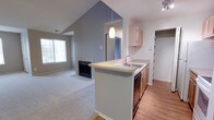 This single-floor residence is designed with an open-concept layout that seamlessly connects the living area and kitchen. The living room is a standout feature, showcasing soaring vaulted ceilings painted in a cool gray-blue tone that lend a sense of spaciousness and airiness to the room. A tiled fireplace with a black glass surround serves as a focal point, adding warmth and character to the space. The carpeted flooring throughout the living area creates a comfortable, soft underfoot feel.

The kitchen is efficiently designed with a wraparound peninsula that doubles as a breakfast bar, topped with a light-toned countertop and equipped with an integrated sink. A decorative pendant light hangs above the peninsula, providing both task and ambient illumination, while a track light fixture adds additional brightness. Light wood cabinetry lines the kitchen walls, complemented by a white refrigerator and a dishwasher. The kitchen features hardwood-style flooring, providing a clean contrast to the carpeted living areas.

A hallway connects the main living spaces to additional rooms, with a window in the living area allowing natural light to fill the space. Overall, the residence maintains a neutral, contemporary palette throughout.