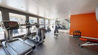Located on E Fort Lowell Road in Tucson, Arizona, this single-room fitness center offers a well-equipped workout space designed to meet a variety of exercise needs. The room features a dark, durable rubber flooring throughout, providing a practical and resilient surface suited for both cardio and strength training activities.

Natural light fills the space through a series of large, dark-framed windows along one wall, offering views of the surrounding outdoor amenity area. A bold orange accent wall adds a dynamic, energizing visual contrast to the otherwise neutral white and gray color palette. A full-length wall mirror on one side of the room amplifies the sense of space and allows users to monitor their form during workouts.

The fitness center is equipped with a range of cardio machines, including treadmills, an elliptical trainer, and a stationary bike. Strength training options include a multi-station cable machine, a weight bench, a dumbbell rack with a varied selection of free weights, kettlebells, and stability balls. A wall-mounted pull-up bar and a chalkboard panel provide additional functional training options.

A wall-mounted split air conditioning unit ensures climate control, and a teal accent door provides a welcoming entry point. The space is thoughtfully arranged to maximize usability while maintaining an open, accessible layout.