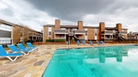 This outdoor community space centers around a generously sized swimming pool with clear turquoise water, designed for leisure and relaxation. The pool is surrounded by an expansive deck constructed from irregularly shaped flagstone pavers in warm tones of tan, gold, and gray, creating a visually striking and durable surface that extends across the entire pool area. Blue lounge chairs are arranged along the poolside, offering ample space for sunbathing and outdoor recreation.

The backdrop of the pool area is defined by a multi-story brick building featuring a classic architectural style with prominent exterior chimneys, dark-trimmed balconies, and exterior staircases. The brick facades convey a sturdy, timeless construction quality consistent throughout the property.

Beyond the pool, a secondary courtyard area is accessed through a wrought iron fence and gate. This space is landscaped with mature trees and features additional flagstone paving that continues the cohesive design of the outdoor areas. A dedicated chemical storage enclosure and utility access are also present within the pool complex, indicating a well-maintained facility. The combination of the distinctive flagstone hardscape, classic brick architecture, and resort-style pool amenities makes this outdoor space a central and functional gathering area for the community.