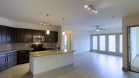 This contemporary single-floor space showcases an open-concept layout that seamlessly connects the kitchen and living area, creating a bright and airy atmosphere. The kitchen is appointed with rich dark cabinetry, gleaming granite countertop surfaces, and a full suite of stainless steel appliances including a range, microwave, and additional cooking appliances. A central island with a built-in sink serves as both a functional workspace and a natural divider between the kitchen and the rest of the living space. Pendant lighting fixtures hang above the island, complementing the modern track lighting installed in the kitchen area. The expansive living area features light-toned hardwood-style flooring that runs throughout, contributing to a clean and cohesive aesthetic. A ceiling fan provides both comfort and a finishing touch to the living space, which benefits from generous natural light streaming through large windows along the far wall. The overall ambiance is modern and welcoming, with a neutral color palette and thoughtful design details that make the space both functional and visually appealing.
