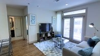 This single-story space features a well-appointed living area designed with a clean, contemporary aesthetic. The living room is characterized by warm hardwood-style flooring that extends throughout, complemented by neutral light gray walls that create a bright and inviting atmosphere. Recessed lighting is integrated into the ceiling alongside a centrally placed ventilation duct, contributing to a polished, modern finish overhead.

Large floor-to-ceiling sliding glass doors and windows flood the space with abundant natural light while providing direct access to an outdoor balcony, offering views of the surrounding area. Sheer white curtains and slatted blinds allow for flexible light control. The living area comfortably accommodates a media console with open shelving, a flat-screen television, and a glass-top coffee table set atop a geometric patterned area rug, anchoring the seating arrangement. Framed artwork adds a decorative touch to the walls, enhancing the overall contemporary feel of the space. A hallway visible in the background suggests additional rooms beyond the main living area, completing this thoughtfully designed single-floor residence.