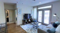This single-story space features a well-appointed living area designed with a clean, contemporary aesthetic. The living room is characterized by warm hardwood-style flooring that extends throughout, complemented by neutral light gray walls that create a bright and inviting atmosphere. Recessed lighting is integrated into the ceiling alongside a centrally placed ventilation duct, contributing to a polished, modern finish overhead.

Large floor-to-ceiling sliding glass doors and windows flood the space with abundant natural light while providing direct access to an outdoor balcony, offering views of the surrounding area. Sheer white curtains and slatted blinds allow for flexible light control. The living area comfortably accommodates a media console with open shelving, a flat-screen television, and a glass-top coffee table set atop a geometric patterned area rug, anchoring the seating arrangement. Framed artwork adds a decorative touch to the walls, enhancing the overall contemporary feel of the space. A hallway visible in the background suggests additional rooms beyond the main living area, completing this thoughtfully designed single-floor residence.