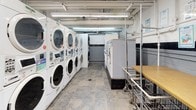 This shared laundry facility is a fully functional, single-floor space designed to accommodate high-volume laundry needs. The room is lined with multiple stacked front-loading washer and dryer units arranged in rows, providing ample capacity for simultaneous use. The machines are modern, commercial-grade appliances equipped with digital controls and coin-operated or card payment systems.

The flooring throughout consists of durable slate-style tile, practical for a high-traffic utility space. Overhead, exposed pipes, ductwork, and fluorescent strip lighting run across the ceiling, delivering bright, even illumination throughout the room. The walls are constructed of painted concrete block, reinforcing the utilitarian character of the space.

A folding area is provided along one wall, featuring a wooden-topped folding table accompanied by wire laundry carts on wheels, making it easy to sort and transport clothing. Informational notices and operating instructions are posted on the walls for user guidance.

Additional amenities include a vending machine, a fire extinguisher, and a wall-mounted clock. A glazed interior door connects the laundry room to an adjacent corridor, ensuring convenient access. The space is straightforward and efficiently organized, prioritizing function and ease of use for its occupants.