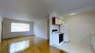 This single-floor space features an open-concept layout that integrates the living area and kitchen into one cohesive environment. The gleaming hardwood floors extend throughout, adding warmth and a polished feel to the interior. The living area is bright and airy, benefiting from a sizable window that invites natural light and offers a view of the surrounding greenery. A baseboard heater runs along the wall beneath the window, providing an efficient heating solution. The kitchen occupies a defined section of the open floor plan, separated by a partial wall. It is equipped with a white electric range and a full-sized white refrigerator, complemented by upper cabinetry in a combination of wood-toned and white finishes, offering practical storage. A ceiling-mounted light fixture provides focused illumination over the kitchen workspace. The walls throughout are finished in a clean, neutral white, creating a bright and versatile backdrop. The overall ambiance is straightforward and functional, with the open layout maximizing the sense of space across the single floor. A doorway visible in the background suggests additional rooms or storage beyond the main living and kitchen area.