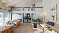 This single-story clubhouse in North Charleston, SC, offers a well-appointed community gathering space defined by modern coastal design and thoughtful amenities. The main living area is expansive and airy, characterized by soaring vaulted ceilings clad in white shiplap paneling that lend a bright, clean aesthetic throughout. Warm wood-look flooring flows seamlessly across the space, complementing the neutral and contemporary furnishings.

A fully equipped fitness center is prominently positioned behind floor-to-ceiling black-framed glass walls and double doors, allowing clear sightlines from the lounge area while maintaining a dedicated workout environment. The lounge itself is furnished with comfortable seating arrangements and accented by curated abstract artwork, creating an inviting atmosphere for socializing or relaxing.

The kitchen and kitchenette area is elegantly finished with soft sage cabinetry, white quartz countertops, and eye-catching herringbone marble-look tile backsplash. Open shelving displays decorative accents, while a beverage cooler and coffee station add convenience. Multiple windows flood the kitchen with natural light and offer direct views of the outdoor pool area, enhancing the overall connection to the surrounding landscape. Small bistro-style tables provide casual seating within this space, making it ideal for community gatherings.