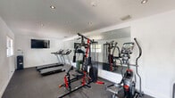 Located at 1931 W 16th Ct in Riviera Beach, Florida, this dedicated fitness center is a well-equipped exercise room designed to support a full range of workout routines. The space features crisp white walls and a clean, bright interior accentuated by recessed ceiling lighting and a wall-mounted flat-screen television for entertainment during workouts.

The exercise room is outfitted with an impressive variety of fitness equipment, including multiple treadmills, elliptical trainers, a multi-function cable strength machine, and additional cardio machines. A set of free weights and dumbbells rounds out the strength training options. The floor is finished with durable rubberized, speckled gym flooring, providing both safety and comfort underfoot.

A large wall-to-wall mirror spans one side of the room, visually expanding the space and allowing users to monitor their form during exercise. Natural light filters in through a window offering a view of the surrounding greenery, while a glass door on one side provides convenient access and additional brightness. A water cooler station is also available for hydration. The space is further equipped with a wall-mounted sanitizing dispenser and waste bin, ensuring cleanliness and convenience throughout. Overall, this fitness center delivers a functional and well-appointed environment for residents seeking an on-site workout facility.