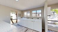 This shared laundry facility is a clean, efficiently designed single-floor space built to accommodate high-volume use. The room is outfitted with a row of full-sized top-load washing machines lined along one wall, complemented by multiple stacked commercial-grade dryers, providing ample capacity for regular laundry needs. A built-in folding counter with a light stone-style surface runs along the washer row, offering a convenient workspace for sorting and folding laundry. The flooring throughout is a light wood-style vinyl plank, lending a clean and modern aesthetic to the utilitarian space. Large sliding windows along the back wall invite generous natural light and offer views of the surrounding outdoor area, including palm trees and adjacent buildings, giving the room an open, airy feel. A skylight further enhances the brightness of the space. The walls are finished in a neutral warm beige tone that keeps the environment feeling tidy and welcoming. A distinctive teal-colored security door at the entrance provides secure access while still allowing light to filter through. The overall design prioritizes functionality and cleanliness, making this a practical and pleasant shared amenity.