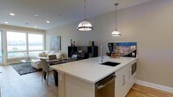 This single-floor space is designed around an open-concept floor plan that seamlessly connects the kitchen, dining, and living areas into one cohesive environment. The kitchen serves as the centerpiece, anchored by a large white island with a smooth quartz-style countertop, an undermount sink with a contemporary arc faucet, and stainless steel appliances including a built-in dishwasher and microwave. Two stylish pendant lights hang above the island, providing focused task lighting while contributing to the modern aesthetic. White cabinetry reinforces the clean, minimalist design throughout the kitchen. The dining area sits adjacent to the kitchen, featuring a dark-toned table with upholstered chairs, creating a functional yet sophisticated space for meals. Beyond the dining area, the living room offers a relaxed atmosphere with a light-colored sofa and a dark modular shelving unit against the neutral gray walls. Warm hardwood flooring flows throughout, adding texture and continuity. A window in the living area brings in natural light, brightening the entire open space and contributing to its airy, welcoming ambiance.