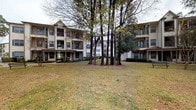 This residential community is composed of several multi-story buildings arranged around shared outdoor spaces, creating a cohesive and well-planned environment. The exterior architecture features a combination of warm brick facades and neutral-toned siding, accented by pitched rooflines, decorative gabled peaks, and private balconies that add both character and functionality to each building.

The community's common areas are a central highlight. Expansive courtyard greenspaces are shaded by a cluster of tall, established trees, giving the grounds a serene, park-like atmosphere. Benches are positioned throughout, offering quiet spots for residents to relax outdoors. Well-maintained landscaping with neatly trimmed shrubs and ground cover lines the building perimeters, adding to the polished appearance of the property.

The pool area serves as a key amenity, featuring a generously sized swimming pool surrounded by a broad brick-paved deck. Multiple lounge chairs and shaded seating areas with umbrella tables provide ample space for outdoor leisure. Lush plantings and mature trees frame the pool area, enhancing privacy and visual appeal.

Overall, the community presents a well-maintained, amenity-rich environment with thoughtful landscaping, comfortable outdoor gathering spaces, and traditional architectural detailing that contribute to a welcoming and comfortable residential atmosphere.