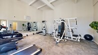 Located at 750 Mobil Ave in Camarillo, CA, this fitness center offers a thoughtfully designed exercise space under soaring vaulted ceilings with exposed white-painted wooden beams, creating an open and airy atmosphere. The exercise room is equipped with a variety of strength and cardio machines, including cable weight machines, a multi-station gym, treadmills, an elliptical, and free weights, all arranged on durable carpet tile flooring. A large wall-mounted mirror spans one side of the room, visually expanding the space and allowing users to monitor their form during workouts. Floor-to-ceiling glass windows along one wall flood the room with abundant natural light and provide views of the lush, tree-lined outdoor surroundings, creating an inviting environment for exercise. An air conditioning unit ensures comfort throughout the year. The facility also includes a dedicated bathroom, finished with a natural stacked-stone accent wall, wood-look tile flooring, and a stone-topped vanity — offering a clean and refined post-workout retreat. The overall design balances functionality with an appealing aesthetic, making this fitness center a well-rounded amenity for its users.