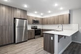This single-floor unit offers a well-organized layout comprising three bedrooms, three full bathrooms, a combined kitchen and living area, a dedicated laundry room, and a connecting hallway. The open-concept kitchen is a standout feature, equipped with stainless steel appliances including a refrigerator, range, and microwave. Flat-panel cabinetry in a warm wood-grain finish lines the walls, complemented by white subway tile backsplash and white quartz countertops. A peninsula with an undermount sink and seating overhang bridges the kitchen and living area, both finished with light wood-look luxury vinyl plank flooring and recessed lighting overhead.

The bedrooms are carpeted and finished in neutral tones, each with windows that bring in natural light and views of the surrounding exterior. One bedroom features a wall-mounted mini-split HVAC unit for independent climate control. The bathrooms are consistent in design, featuring wood-grain vanities with white countertops, large mirrors, and tub-shower combinations with white tile surrounds and light wood-look flooring. The dedicated laundry room houses a full-size front-loading washer and dryer alongside the water heater, finished with the same vinyl plank flooring found throughout the common areas.
