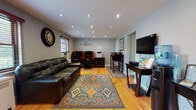 This well-appointed single-floor space is designed to serve multiple purposes with a practical and welcoming layout. The main area features warm hardwood flooring throughout, soft neutral gray walls, and recessed lighting that provides even, consistent illumination across the space.

The lounge area is anchored by a full-sized leather sofa set atop a colorful geometric area rug, offering a comfortable place to relax. Multiple windows dressed with horizontal blinds allow natural light to filter in, enhancing the open feel of the room. A radiator beneath each window provides reliable heating.

Adjacent to the lounge, a dedicated workstation area includes two rolltop-style computer desks with ergonomic chairs and a multi-function printer, making it well-suited for administrative or office tasks. A high-top table with bar stools provides an additional surface for dining or collaborative work, positioned near a wall-mounted flat-screen television and a console table with media equipment.

A compact kitchenette tucked into one corner is efficiently outfitted with rich wood cabinetry, a granite-style countertop, an undermount sink, a microwave, a toaster, and a stainless steel mini-refrigerator — ideal for light meal preparation. A doorway with French glass panels leads to an additional room, further extending the utility of the overall space.