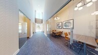 Located at 3240 W Harmon Ave in Las Vegas, NV, this single-floor professional suite offers a thoughtfully designed layout encompassing multiple meeting and conference rooms. The space is defined by its striking architectural features, most notably the soaring double-height ceilings and expansive floor-to-ceiling windows that flood the corridor and rooms with abundant natural light. A central hallway connects the individual meeting rooms, each enclosed by sleek, full-height glass partition walls with sliding glass doors, creating a sense of openness while maintaining acoustic separation. The patterned blue-toned carpeting throughout adds a sophisticated, cohesive visual element to the space. Each meeting room is outfitted with a conference table, ergonomic chairs, and contemporary globe pendant lighting. Select rooms are equipped with wall-mounted displays for presentations, while framed abstract artwork decorates the walls, contributing to a polished, professional ambiance. Two smaller private meeting rooms are positioned side by side and share a similar intimate configuration, while a larger conference room accommodates more attendees around a central table. Large exterior-facing windows along the corridor provide views of the surrounding area, enhancing the bright and welcoming atmosphere of this well-appointed professional space.