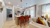 This single-story residence offers a thoughtfully designed open-concept layout that seamlessly connects the kitchen, dining, and living areas. The kitchen is a standout feature, outfitted with flat-front wood-tone cabinetry extending to the ceiling, stainless steel appliances including a refrigerator and microwave, and a white countertop island with bar seating for three. Pendant lighting above the island and recessed ceiling lights enhance the bright, contemporary atmosphere. Light wood-look flooring runs throughout the main living areas, contributing to a clean, cohesive aesthetic. A door with adjacent windows in the kitchen provides natural light and exterior access.

The space includes two bedrooms, both carpeted and outfitted with ceiling fans and windows dressed with patterned drapes. The primary bedroom features a tray-style ceiling accent and ample space for a full furniture arrangement. A full bathroom includes a wood-tone vanity with white countertop, a soaking tub with shower combo, and matching light wood flooring. A dedicated laundry area accommodates a stacked full-size washer and dryer alongside shelving and hanging storage, adding practical functionality to the overall layout.