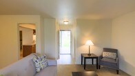 This single-floor space features a thoughtfully arranged layout that connects the main living area to adjacent rooms with ease. The living room provides a comfortable gathering space, furnished with a plush sofa accented by patterned throw pillows and complemented by a dark wood side table topped with a warm table lamp, adding a cozy ambiance to the room. Neutral-toned carpet grounds the living area, while light-colored walls reflect a clean, understated aesthetic throughout.

A hallway leads directly to an exterior door, which allows natural light to pour into the interior and offers views of an outdoor green space beyond, creating a welcoming sense of openness. The transition between spaces is smooth and unobstructed, contributing to the overall functional flow of the floor plan.

Visible to the side of the living area, the kitchen features wood-toned cabinetry and a countertop workspace, indicating a practical cooking space within close proximity to the main living zone. Flush-mount ceiling lighting illuminates both the hallway and living area uniformly. The overall ambiance is bright, neutral, and practical, making the space well-suited for everyday living.