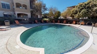 This outdoor amenity area centers around a generously sized swimming pool, available in both a freeform organic shape and a more traditional rectangular layout depending on the specific complex. The pool deck is finished in light-toned concrete, providing ample space for sunbathing and relaxation. Multiple lounge chairs and side tables are arranged around the perimeter, offering residents comfortable spots to enjoy the outdoors. A wrought iron perimeter fence encloses the space, ensuring security while maintaining open sightlines to the surrounding mature trees and landscaping, which provide natural shade throughout the day. An adjacent spa or hot tub is visible in one view, adding a wellness-oriented feature to the amenity space. A dedicated pool house structure, finished in stucco with dark wood-trimmed doorways and a wall-mounted exterior light, provides direct access to the pool deck through glass-paneled doors. The interior of the pool house is a compact utility space with concrete flooring. Posted pool rules signage is visible near the entrance. The multi-story residential buildings surrounding the pool area feature balconies that overlook the amenity space, reinforcing the sense of a well-integrated community environment bathed in abundant natural sunlight.