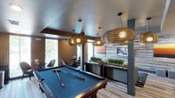 This single-floor recreational space in Little Elm is thoughtfully designed to blend comfort and entertainment in a modern, inviting atmosphere. The focal point of the space is a full-sized billiards table, set beneath a striking arrangement of woven rattan pendant lights that cast a warm, ambient glow throughout the room. The light wood-plank flooring adds a clean, contemporary foundation that complements the earthy tones of the décor. Large windows along one wall flood the interior with natural light and offer views of the surrounding greenery, creating a bright and airy feel. A dedicated lounge area with stylish seating provides a relaxed setting for socializing. Along the back wall, a horizontal shiplap-style accent wall in muted tones serves as a backdrop for a flat-screen display, flanked by decorative greenery arrangements and dark wood credenzas that add a refined touch. Structural columns are incorporated seamlessly into the design, enhancing the architectural character of the space. The textured ceiling adds depth overhead, while the overall palette of warm neutrals, natural wood, and soft lighting contributes to a welcoming and upscale ambiance well-suited for leisure and community gathering.