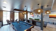 This single-floor recreational space in Little Elm is thoughtfully designed to blend comfort and entertainment in a modern, inviting atmosphere. The focal point of the space is a full-sized billiards table, set beneath a striking arrangement of woven rattan pendant lights that cast a warm, ambient glow throughout the room. The light wood-plank flooring adds a clean, contemporary foundation that complements the earthy tones of the décor. Large windows along one wall flood the interior with natural light and offer views of the surrounding greenery, creating a bright and airy feel. A dedicated lounge area with stylish seating provides a relaxed setting for socializing. Along the back wall, a horizontal shiplap-style accent wall in muted tones serves as a backdrop for a flat-screen display, flanked by decorative greenery arrangements and dark wood credenzas that add a refined touch. Structural columns are incorporated seamlessly into the design, enhancing the architectural character of the space. The textured ceiling adds depth overhead, while the overall palette of warm neutrals, natural wood, and soft lighting contributes to a welcoming and upscale ambiance well-suited for leisure and community gathering.
