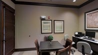 This single-floor space presents a well-appointed interior designed to balance comfort and functionality. The main area centers around a dark wood square dining table surrounded by brown upholstered chairs, creating an intimate setting suitable for small gatherings or meetings. A small decorative green plant serves as a tasteful centerpiece on the table, adding a touch of natural warmth to the setting.

The walls are finished in a soft, neutral beige tone, contributing to the room's calm and understated ambiance. Two framed landscape-style artworks with muted, earthy tones are displayed side by side, enhancing the visual appeal of the space without overwhelming it. Dark wood crown molding runs along the upper perimeter of the walls, providing a refined architectural detail that anchors the room's design.

Recessed lighting overhead ensures the space is well-lit, while a fire safety device is mounted on the wall, reflecting attention to safety compliance. To one side, a workstation with a computer monitor and a dark cabinetry unit adds a practical element, suggesting the space serves a dual purpose as both a gathering and work area.