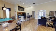 This spacious clubroom is designed for comfort and versatility, offering a welcoming environment for social gatherings and community events. The space is unified by warm hardwood-style flooring and soaring ceilings that create an open, airy atmosphere throughout.

At the heart of the room is a fully equipped kitchen, outfitted with warm wood cabinetry, dark granite countertops, and black appliances including a range, microwave, refrigerator, and dishwasher. A raised bar-height island with seating provides a casual dining option, while pendant lighting adds a refined touch overhead. Crown molding and recessed lighting further elevate the kitchen area.

The lounge area is furnished with plush navy blue sofas and armchairs, centered around a flat-screen television and a media console with a built-in electric fireplace. Decorative wall art and accent pillows contribute to the polished, inviting aesthetic.

A dedicated dining area features a round table with upholstered chairs, positioned near large arched windows dressed with valances that allow abundant natural light to filter in. French doors open directly to an outdoor patio, enhancing the connection between interior and exterior spaces. The overall design strikes a balance between functionality and style, making this clubroom a versatile and comfortable community hub.